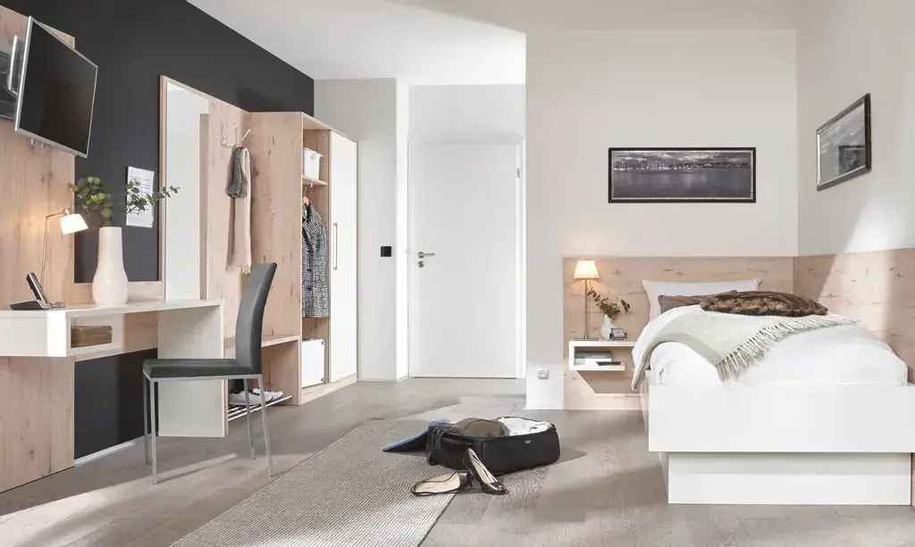 Weisses Bett mit Nachtkonsole rechts, links Schreibtisch mit Stuhl und Garderobe, geoeffneter Kleiderschrank und Koffer am Boden